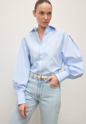 Lyseblå skjorte med knapper og pufærmer, kombineret med lyse, højtaljede jeans i denim. Hvidt bælte med spænde i guld fuldender looket.