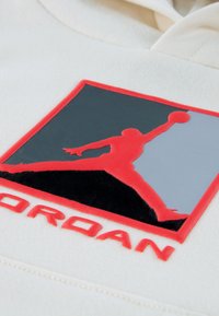 Φούτερ με κουκούλα σε κρεμ απόχρωση, με μαύρο και κόκκιν ο λογότυπο Jumpman με λαμπερή υφή, περιγραμμένο με κόκκινο, και τη λέξη "JORDAN" με μικρά κόκκινα γράμματα.