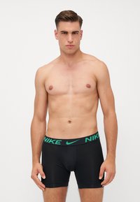 Sort sportsbokser med en grøn Nike-logo på waistbande. Materialet ser glat og tætsiddende ud, med en længde til midt på låret og ingen synlige mønstre.