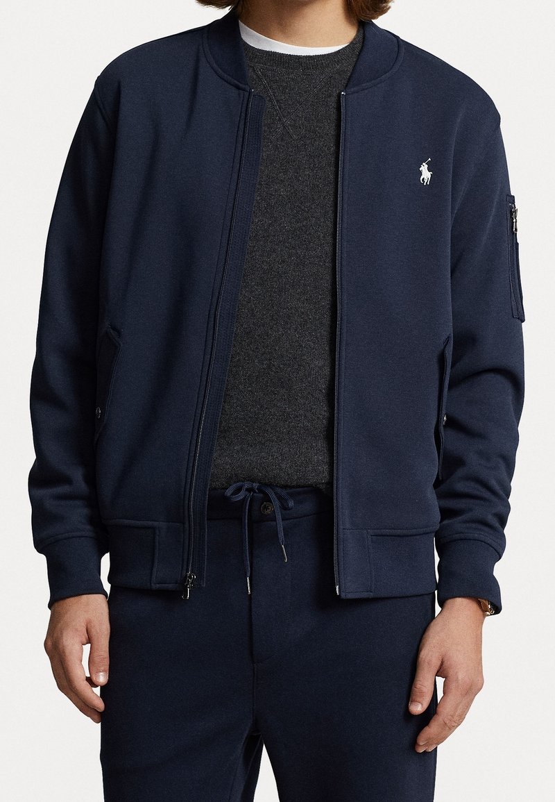Homme portant une veste bomber zippée bleu marine sur un pull gris foncé et un pantalon bleu marine assorti avec cordon de serrage, avec un petit logo blanc sur la poitrine de la veste.