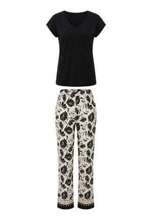 Mujer de pie descalza con una camiseta negra de manga corta con cuello en V y pantalones crema con estampado floral negro y puños estampados.