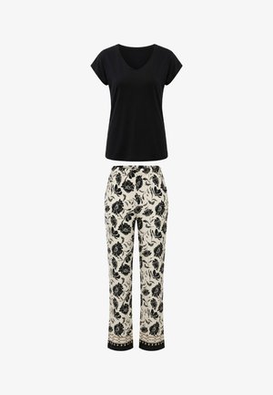 Mujer de pie descalza con una camiseta negra de manga corta con cuello en V y pantalones crema con estampado floral negro y puños estampados.