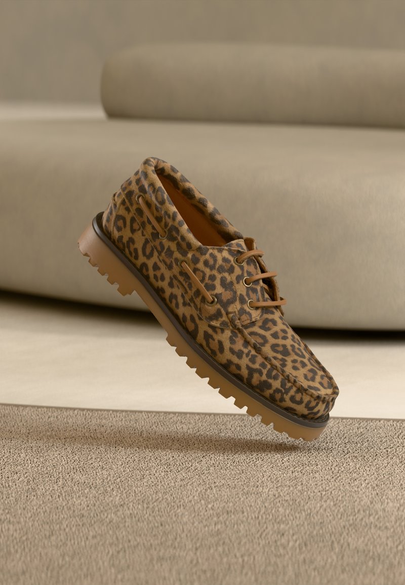 Mocassins à imprimé léopard marron et noir, semelle épaisse en caoutchouc beige, et lacets marron, posés sur un tapis beige devant un canapé beige.