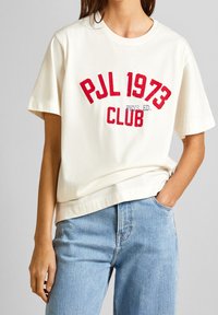 Krämfärgad bomulls-T-shirt med röd grafisk text "PJL 1973 CLUB" och "PHYS. ED." på framsidan. Korta ärmar, avslappnad passform, kombinerad med ljusblå jeans.