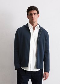 Navy zip-up hoodie with a soft texture, featuring a hood and front zip closure, worn over a white button-up shirt.