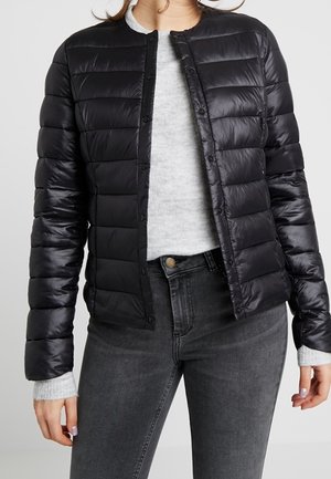 Femme portant une veste matelassée noire à boutons par-dessus un pull gris clair et un jean gris foncé, debout les mains détendues le long du corps.