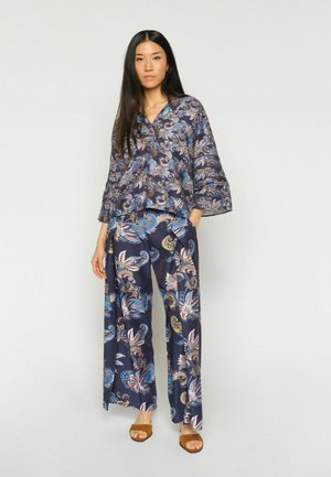 Woman standing wearing matching loose floral print blouse and wide-leg pants with brown open-toe sandals against plain background.