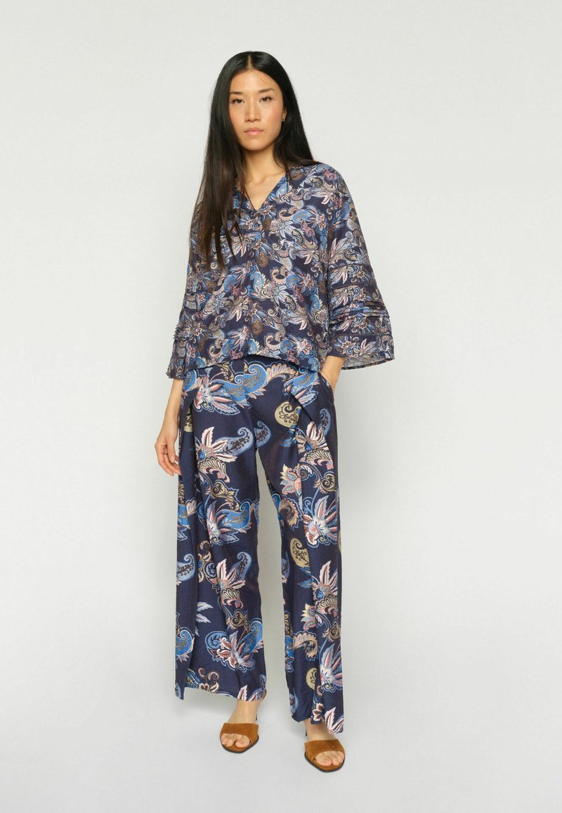 Femme debout portant un chemisier ample assorti à motif floral et un pantalon à jambes larges avec des sandales marron à bouts ouverts, sur fond uni.