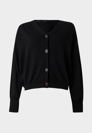 Femme debout, une main dans la poche, portant un cardigan noir boutonné, un haut noir, un pantalon blanc à larges jambes et des chaussures noires.