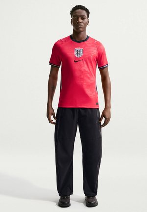 Uomo in piedi che indossa una maglia da calcio dell'Inghilterra rossa con emblema, pantaloni neri Nike e sneakers nere contro uno sfondo semplice.
