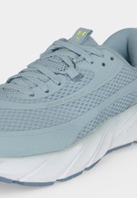 Ανοιχτό μπλε αθλητικό παπούτσι Under Armour με πλέγμα στο επάνω μέρος, επενδεδυμένο γιακά και λευκή μαλακή σόλα που δείχνει λεπτομερείς κορδόνια και το λογότυπο της μάρκας στη γλώσσα.