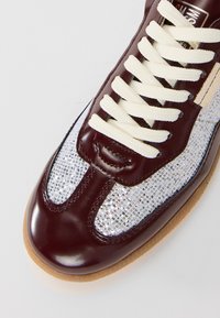 Burgundska usnjena teniska obutev z belimi ravnimi vezalkami, srebrnimi bleščečimi tkaninskimi paneli in gumijastim podplatom, prikazana z zgornje sprednje strani pod kotom.