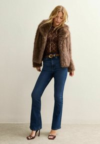 Veste en fausse fourrure marron, haut en dentelle, jeans peints bleu et talons hauts marron. La tenue présente une taille ceinturée et un long collier pendentif.