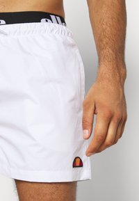 Weiße Badehose mit elastischem Bund, der ein schwarzes Logo zeigt. Bemerkenswerte Stickerei in Rot, Gelb und Schwarz am Saum.