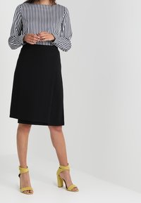 Femme debout portant une jupe noire à mi-mollet, un haut rayé noir et blanc à manches longues, et des sandales à talons hauts jaunes.