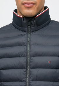 Ναυτικό μπουφάν με κατακόρυφο σχεδιασμό καπιτονέ, ψηλό γιακά με κόκκινες και λευκές ρίγες και μπροστινό φερμουάρ. Μικρό λογότυπο στο στήθος.