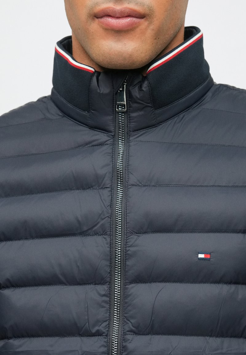 Marineblaue Pufferjacke mit horizontalem Steppdesign, hohem Kragen mit roten und weißen Streifen und einem Frontreißverschluss. Kleines Logo auf der Brust.