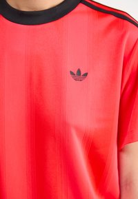 Raudona sportinė marškinė su juodu apykaklės kraštu, vertikaliomis linijomis ir mažuoju juodu "Adidas" logotipu ant krūtinės. Pagaminta iš lygaus, lengvo audinio.