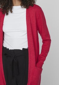 Cárdigan rojo con una textura de punto, diseño de frente abierto y puños acanalados. Combinado con una camiseta blanca y pantalones negros de cintura alta.