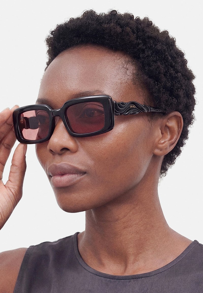 Gros plan d'une femme aux cheveux courts et bouclés portant de grandes lunettes de soleil noires carrées avec des verres rouge foncé et un haut sombre sans manches.