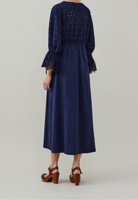 Sininen maksimekko yksityiskohtaisilla broderie-hihnoilla, joustava vyötärö ja sivuaukot. Pukeutuneena ruskeisiin tasan korkoisiin sandaaleihin, joissa on texturoitu muotoilu.