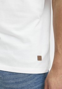 Tricou alb din bumbac cu mâneci scurte, având o etichetă pătrată din piele maro cusută în marginea din stânga jos. Sunt vizibile blugi bleu deschis.