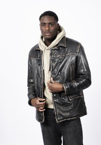 Chaqueta de cuero negra con un delantero abotonado, bolsillos con parche y un acabado texturizado. Usada sobre una sudadera ligera de color beige, combinada con jeans oscuros.