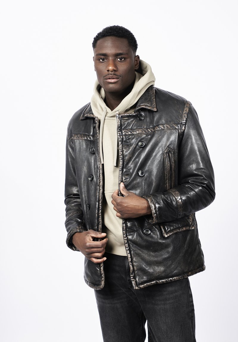 Chaqueta de cuero negra con un delantero abotonado, bolsillos con parche y un acabado texturizado. Usada sobre una sudadera ligera de color beige, combinada con jeans oscuros.