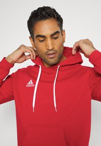 Roter Baumwoll-Hoodie mit weißen Kordeln und einer Kängurutasche. Mit dem weißen Adidas-Logo auf der linken Brustseite. Glatte Textur.