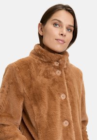 Brun faux fur frakke med høj krave, der har tre lysebrune knapper, blød tekstur og enkelt design. Velegnet til afslappet brug.