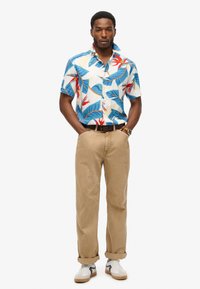 Kortærmet button-up skjorte i cremefarvet med blå, grønne og røde bladmønstre, parret med beige bukser og hvide sneakers med grå detaljer.