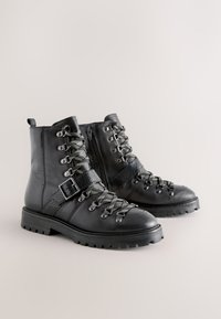 Musta nahkainen nilkkurit, joissa on teksturoitu paneeli, hopeiset metalliosat ja nauhakiinnitys; sisältävät sivuvetoketjun ja rouhean pohjan.