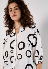 White button-up shirt featuring a black circular pattern. The fabric has a light texture, with three-quarter sleeves and a collar.