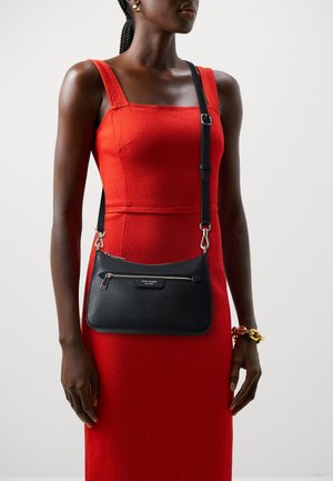 Black leather crossbody bag with textured finish, a zippered front pocket, and a detachable strap, held against a red dress backdrop.
