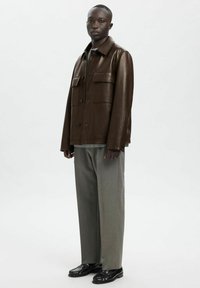 Rjava usnjena jakna z dvema sprednjima žepoma, zapiranjem na gumbe in ovratnikom. Kombinirana s svetlo sivimi hlačami in črnimi čevlji.