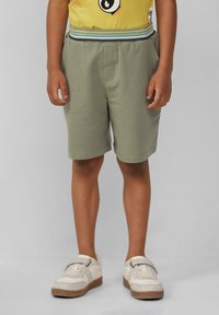 Pantaloni corti di lunghezza al ginocchio, di colore verde oliva con una trama morbida, con vita elastica e strisce blu e gialle. Abbinati a sneaker bianche.