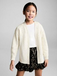 Witte gebreide cardigan met een gestructureerd patroon, met een open voorkant, gedragen over een korte zwarte rok met gouden stippen en ruches.