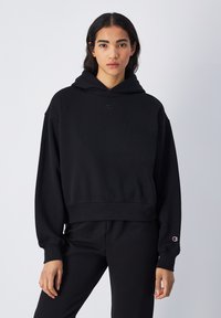 Sweat à capuche noir en tissu doux, avec un design court, une coupe oversized et un petit logo sur la poitrine et la manche.