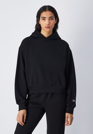 Sweat à capuche noir en tissu doux, avec un design court, une coupe oversized et un petit logo sur la poitrine et la manche.
