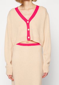 Lyhennetty beige villatakki, jossa on V-pääntie ja kirkkaan vaaleanpunaiset yksityiskohdat. Takissa on kolme nappia ja pitkät hihat. Pehmeä neulepinta ja istuva malli.