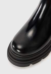 Bottine noire à talon avec une finition en cuir brillant. Présente un bout arrondi et une semelle en caoutchouc noir texturé. Pas de matériel ou d'accents visibles.