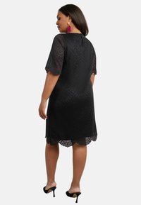 Černé krajkové šaty s krátkými krajkovými rukávy, vlnitým lemem a detaily v oblasti zad. Doplněno růžovými výraznými náušnicemi a černými lodičkami.