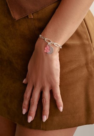 Silver chain bracelet featuring a pink gummy bear charm and a circular pendant. The hand rests on a brown suede surface.