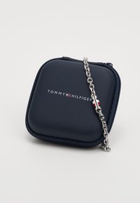 Dunkelblaues Etui mit Reißverschluss und silberner Kette, mit dem "TOMMY HILFIGER"-Logo in Weiß auf der Vorderseite, glatte Textur.