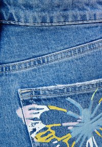 Blå denimtyg med en sliten ficka som har djärva, färgglada abstrakta målningsmönster i gult, rosa och blått.