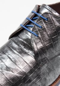 Chaussure noire en cuir à lacets avec une texture en motif crocodile, dotée de lacets bleus et d'accents de semelle marron. Vue rapprochée du design supérieur.