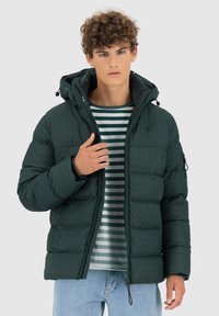 Giacca a puffo verde con zip frontale, cappuccio e polsini elasticizzati. Indossata sopra una maglietta a righe. Presenta una texture cucita e tasche laterali.