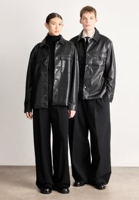 Divas personas nēsā melnas mākslīgās ādas jakas ar pogu aizdari un krūšu kabatām, ko papildina melnas plašu kāju bikses, fonā ar neitrālu krāsu.