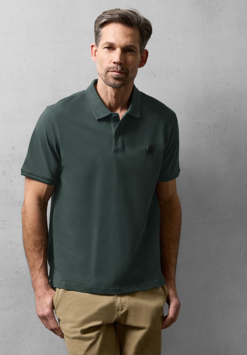 Homme portant un polo vert foncé et un pantalon beige, debout contre un mur gris uni, les mains dans les poches.