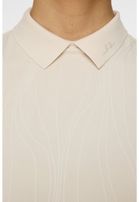 Lähikuva henkilöstä, joka on pukeutunut vaaleanbeigeen kauluspaitaan, jossa on hienovaraisia, tekstuurisia aalto-kuvioita ja pieni kohopainettu logo kauluksessa.